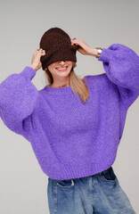 Happy, fashionable blonde woman wearing trendy, stylish violet sweater and winter cap, posing in studio with toothy smile.