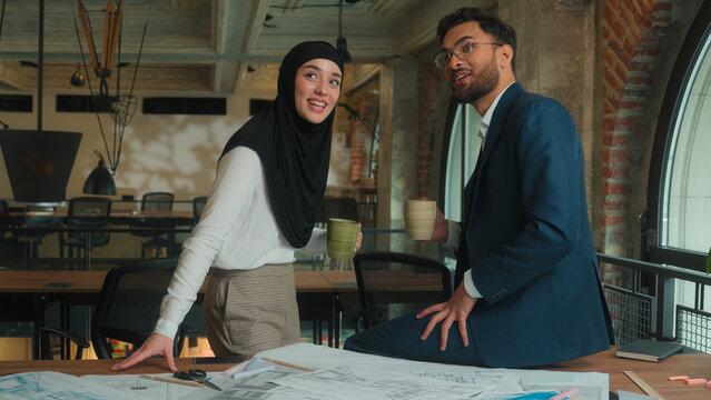 Two architects contractors engineers colleagues architecture project Arabian muslim designers man woman in hijab discuss blueprint draft of office building businessman businesswoman drink coffee smile - Powered by Adobe