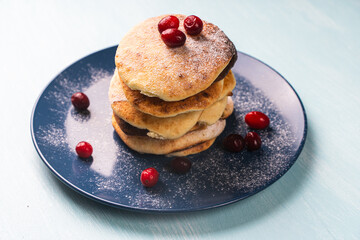 Cottage cheese pancakes with cranberries are stacked on a blue plate on a light blue table, sprinkled with powdered sugar
