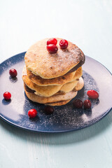 Cottage cheese pancakes with cranberries are stacked on a blue plate on a light blue table, sprinkled with powdered sugar
