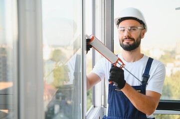 Young man, dressed in a uniform, doing repair in the house. Handyman is applying silicone from a...