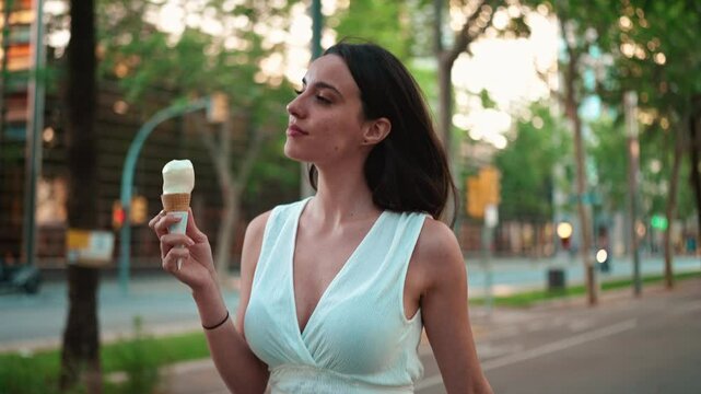 Beautiful woman with freckles and dark loose hair wearing white top is walking down the street and testing ice cream. Cute girl enjoys ice cream on modern city background