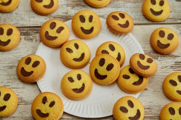 Yellow cookies in the shape of smiling emojis arranged on a wooden table with a white plate in close-up, good mood, friendliness and warmth, good family atmosphere, happiness and joy.