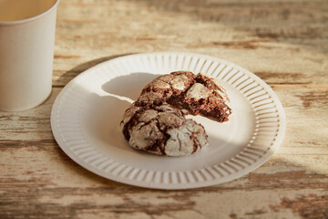 A plate with a broken half of a chocolate cookie with cracks on an old rustic wooden table in a beam of sunlight, with a disposable cup nearby, close-up, homemade baking, rustic breakfast.