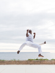 Capoeira martial artist executing a high kick while wearing white, alongside the ocean outdoors
