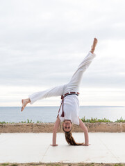 A woman practicing capoeira handstand outside by the sea, showcasing balance and fitness