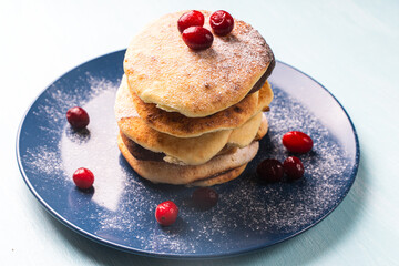 Cottage cheese pancakes with cranberries are stacked on a blue plate on a light blue table, sprinkled with powdered sugar