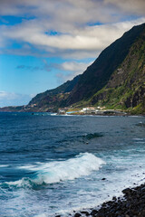 Powerful waves crashing against Madeira’s rocky shoreline, with steep green cliffs and the vast Atlantic Ocean under a partly cloudy sky.