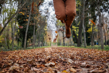 Legs of a person jumping over fallen orange leaves in an autumn park, dressed in matching warm-toned clothing. A dynamic and seasonal scene capturing the joy and energy of fall