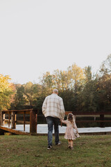 Fototapeta premium Father and Daughter Walking Hand-in-Hand by a Rustic Dock in Autumn – Heartwarming Family Moment, Scenic Nature Background, Bonding Time, Peaceful Outdoor Setting, Warm Light Capturing Parent-Child