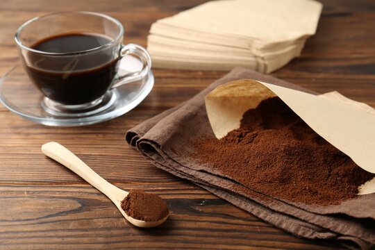 Paper filter, ground coffee and cup of drink on wooden table, closeup
