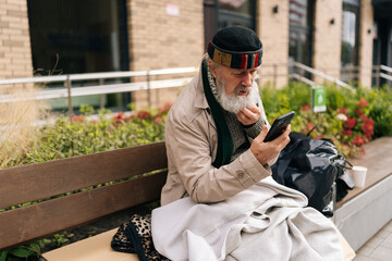 Pensive retired beggar man wrapped in warm blanket using smartphone sitting on urban bench outside building on cold autumn day. Concept of poverty and vulnerability, evoking empathy and compassion.