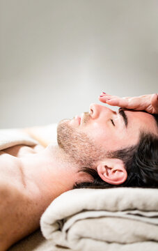 Vertical photo of an adult bearded man in his 30s lying in a spa getting a facial massage on his forehead. Concept of attractive men getting facial massages. Copyspace for advertising.