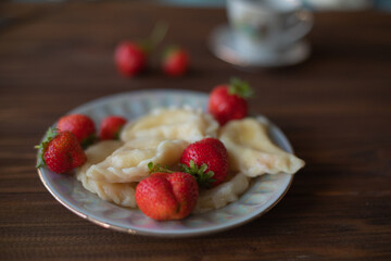 A plate of dumplings with strawberries served on a wooden table, dusted with powdered sugar. A cup of tea in a vintage porcelain cup adds a touch of elegance to this homemade meal.
