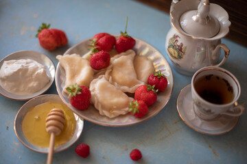 A plate of sweet dumplings with strawberries on a light blue table, with a bite taken out to reveal the creamy filling. A cup of tea sits nearby, completing the relaxing scene.
