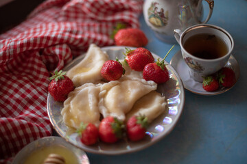 A beautifully styled table setup with a plate of dumplings and fresh strawberries, accompanied by a cup of tea, honey, and a vintage teapot. The red gingham cloth adds a charming rustic touch against 
