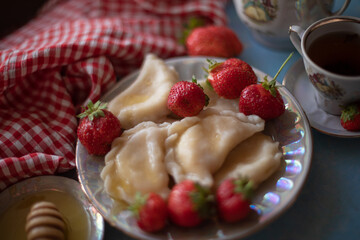 A beautifully styled table setup with a plate of dumplings and fresh strawberries, accompanied by a cup of tea, honey, and a vintage teapot. The red gingham cloth adds a charming rustic touch against 