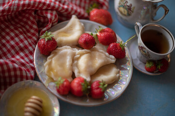A beautifully styled table setup with a plate of dumplings and fresh strawberries, accompanied by a cup of tea, honey, and a vintage teapot. The red gingham cloth adds a charming rustic touch against 