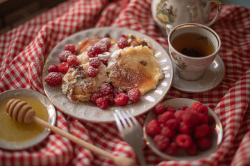 Golden-brown cottage cheese pancakes served with fresh raspberries, lightly dusted with powdered sugar. The plate is accompanied by a vintage teapot, a cup of tea, and a red gingham cloth, creating a 