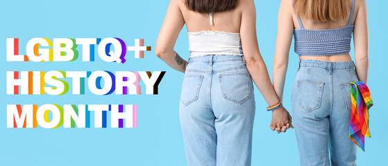 Young lesbian couple holding hands with LGBT flag in jeans pocket on blue background, back view