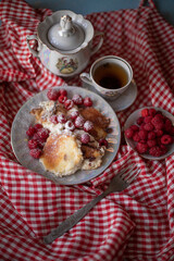Golden-brown cottage cheese pancakes served with fresh raspberries, lightly dusted with powdered sugar. The plate is accompanied by a vintage teapot, a cup of tea, and a red gingham cloth, creating a 