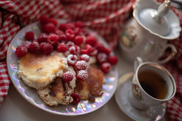 Golden-brown cottage cheese pancakes served with fresh raspberries, lightly dusted with powdered sugar. The plate is accompanied by a vintage teapot, a cup of tea, and a red gingham cloth, creating a 