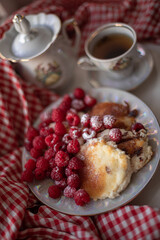 Golden-brown cottage cheese pancakes served with fresh raspberries, lightly dusted with powdered sugar. The plate is accompanied by a vintage teapot, a cup of tea, and a red gingham cloth, creating a 