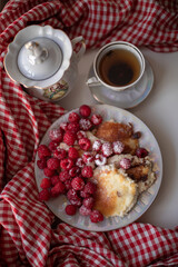 Golden-brown cottage cheese pancakes served with fresh raspberries, lightly dusted with powdered sugar. The plate is accompanied by a vintage teapot, a cup of tea, and a red gingham cloth, creating a 