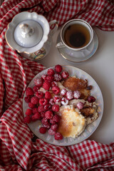 Golden-brown cottage cheese pancakes served with fresh raspberries, lightly dusted with powdered sugar. The plate is accompanied by a vintage teapot, a cup of tea, and a red gingham cloth, creating a 