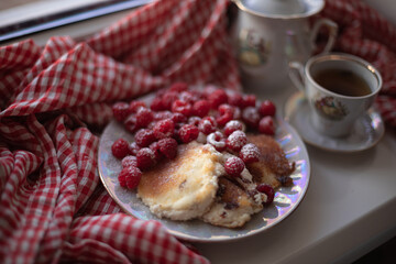 Golden-brown cottage cheese pancakes served with fresh raspberries, lightly dusted with powdered sugar. The plate is accompanied by a vintage teapot, a cup of tea, and a red gingham cloth, creating a 