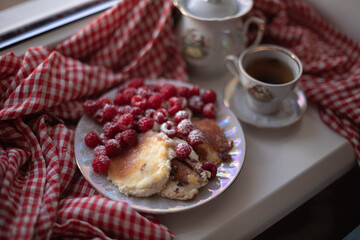 Golden-brown cottage cheese pancakes served with fresh raspberries, lightly dusted with powdered sugar. The plate is accompanied by a vintage teapot, a cup of tea, and a red gingham cloth, creating a 