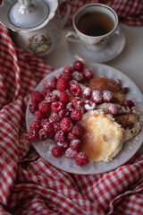Golden-brown cottage cheese pancakes served with fresh raspberries, lightly dusted with powdered sugar. The plate is accompanied by a vintage teapot, a cup of tea, and a red gingham cloth, creating a 
