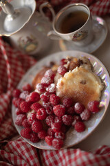 Golden-brown cottage cheese pancakes served with fresh raspberries, lightly dusted with powdered sugar. The plate is accompanied by a vintage teapot, a cup of tea, and a red gingham cloth, creating a 