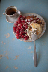 Golden-brown cottage cheese pancakes served with fresh raspberries, lightly dusted with powdered sugar. The plate is accompanied by a vintage teapot, a cup of tea, and a red gingham cloth, creating a 
