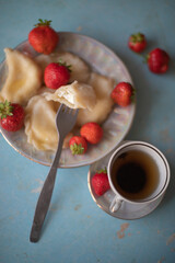 A plate of sweet dumplings with strawberries on a light blue table, with a bite taken out to reveal the creamy filling. A cup of tea sits nearby, completing the relaxing scene.
