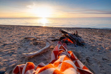 A peaceful sunset view over a calm beach with a bonfire and a blanket in the foreground