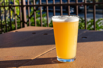 A tall cold glass of hazy IPA beer with froth on top. The clear pint glass of ale is on a brown table with a metal fence in the background. The liquid is a bright yellow color with the sun shining.