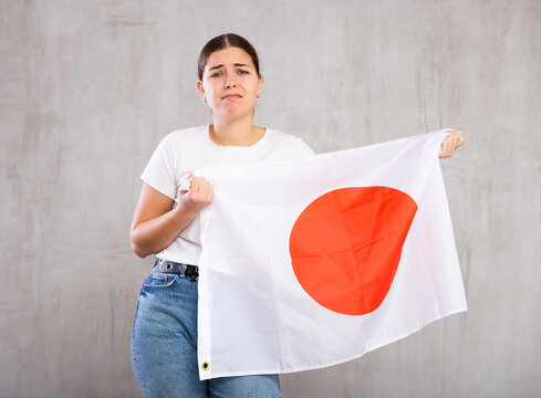 Disappointed young woman in casual clothes with state flag of Japan against gray wall background indoors