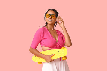 Young African-American woman with skateboard listening to music on pink background