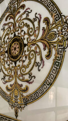 Luxurious ornate golden design with intricate floral and scrollwork details on a white marble surface. Features a symmetrical pattern surrounded by Greek key borders, exuding elegance and timeless bea