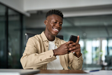 Happy busy Black business man employee using cellphone at work. Smiling young adult African male worker holding mobile phone smart technology gadget in hands working in office sitting at desk.