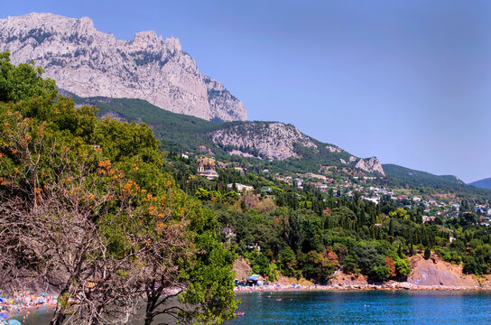 Scenic summer view of a resort mountain coastline with a beach, lush greenery, clear blue sea and wooded hills under a sunny sky.