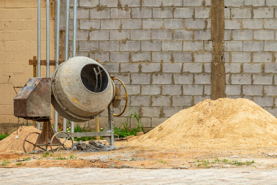 Uma betoneira com um monte de areia ao lado, e um muro de tijolos de concreto ao fundo. Um canteiro de obras.