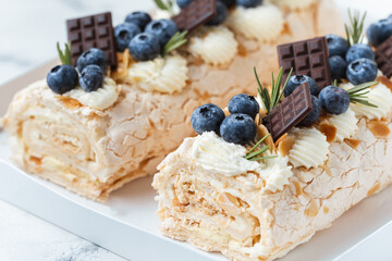 Homemade meringue roll decorated with almond flakes, blueberries and mini chocolate bars in the white gift box on the white background. Meringue roulade with whipped cream on top.