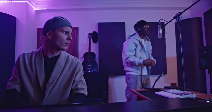 Two music artists collaborate in a professional recording studio, illuminated with neon lights, creating music.