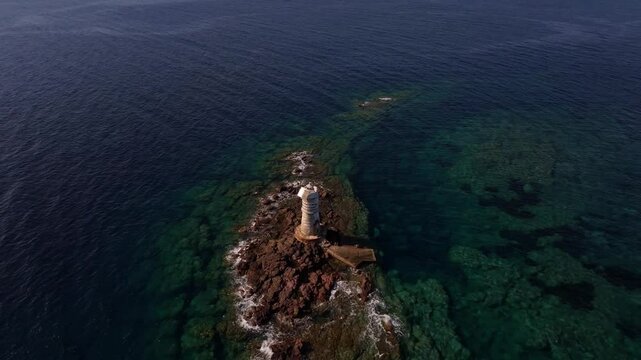 Drone captures Faro Mangiabarche, white lighthouse contrasts with blue waters and brown rocks under the Sardinian sun.