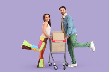 Happy young couple with shopping cart of cardboard boxes on purple background