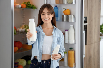 Young woman with bottles of yoghurt near open fridge in kitchen