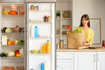 Young woman with paper bag of food on counter near open fridge in kitchen