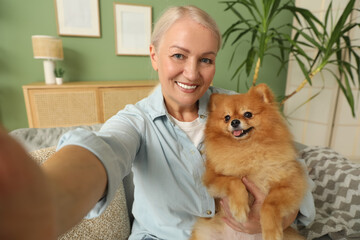 Happy senior woman with cute Pomeranian dog taking selfie on sofa at home